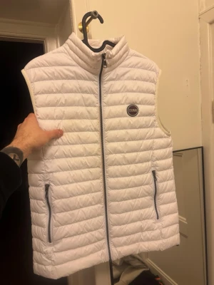 Vit quiltad dunväst från Colmar - Snygg vit dunväst från Colmar med quiltad design och hög krage. Västen har dragkedja framtill och två fickor med dragkedja. Perfekt att slänga över en hoodie eller tröja för extra värme. Klassisk och stilren look. Använd max 3 gånger