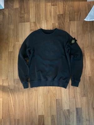 Svart sweatshirt från Stone Island - Säljer en svart sweatshirt från Stone Island med stort ton-i-ton broderat logomärke på bröstet och den klassiska patchen på vänster ärm. Tröjan har rund halsringning, ribbade muddar och är gjord i mjuk bomull. Perfekt för en clean och stilren look. Obs! Två små hål ( bild finns )