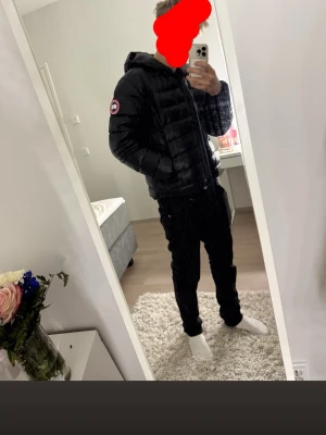 Svart pufferjacka från Canada Goose - Snygg svart pufferjacka från Canada Goose med huva och dragkedja. Jackan har det klassiska Canada Goose-märket på ärmen och är tillverkad i ett glansigt material som ger en modern look. Perfekt för kalla dagar när du vill hålla stilen.