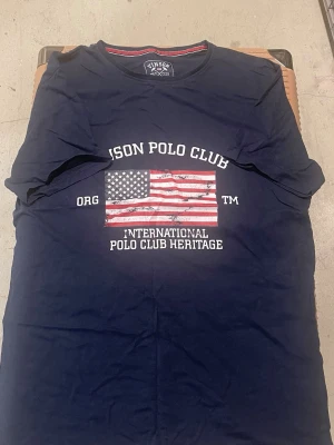 Marinblå t-shirt från Vinson Polo Club - Snygg marinblå t-shirt från Vinson Polo Club med tryck av amerikanska flaggan och texten 'International Polo Club Heritage' på bröstet. Klassisk rund halsringning och korta ärmar. Perfekt för en avslappnad och sportig stil.