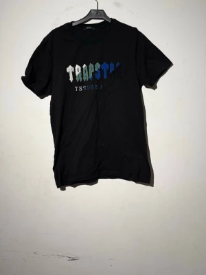 Svart TRAPSTAR t-shirt med tryck - Säljer en svart t-shirt från Trapstar med färgglatt tryck på bröstet där det står 'TRAPSTAR'. T-shirten har rund halsringning och korta ärmar. Perfekt för dig som gillar streetwear och vill ha en stilren men ändå iögonfallande look.