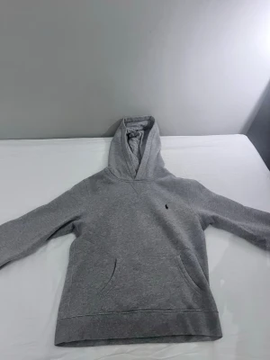 Grå hoodie från Polo Ralph Lauren - Klassisk grå hoodie från Polo Ralph Lauren med broderad logga på bröstet. Hoodien har känguruficka framtill, ribbade muddar och är tillverkad i mjuk bomullsblandning. Perfekt för en avslappnad och stilren look.
