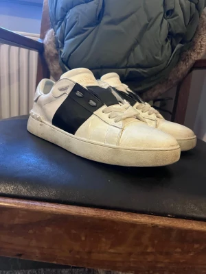 Valentino Garavani Open sneakers - Säljer ett par Valentino Garavani Open sneakers i vitt skinn med bred svart läderdetalj över snörningen och ikoniska nitar på hälen. Skorna har rund tå, platt sula och klassisk snörning. Perfekt för dig som gillar lyxiga och stilrena sneakers med edgy detaljer.