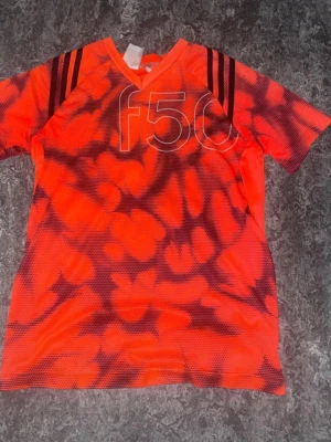 Adidas f50 röd sporttröja - Cool röd sporttröja från Adidas med svart mönster och f50-tryck på bröstet. Tröjan har V-ringning och tre svarta ränder på axlarna. Perfekt för träning eller fotboll. Tillverkad i lätt och ventilerande material.