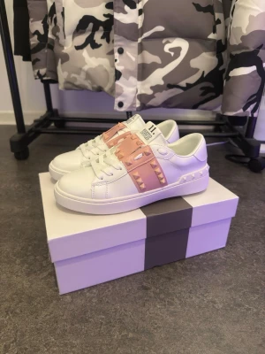  Valentino sneakers  - Snygga vita sneakers från Valentino med rosa band och pyramidnitar på sidan. Skorna har klassisk snörning och är tillverkade i skinn för en lyxig känsla. Perfekta för dig som vill ha något extra till din outfit.