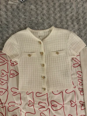 Vit kortärmad kofta från H&M - Snygg vit stickad kofta från H&M med korta ärmar och guldiga knappar framtill. Koftan har två dekorativa fickor på bröstet och en rak, croppad passform. Använd ett fåtal gånger.