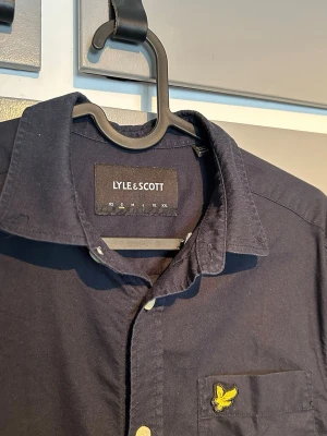 Mörkblå skjorta från Lyle & Scott - Klassisk mörkblå skjorta från Lyle & Scott med gul broderad logga på bröstfickan. Skjortan har långa ärmar, vita knappar och en stilren, tidlös design i mjuk bomull. Perfekt för dig som gillar enkel men snygg stil.