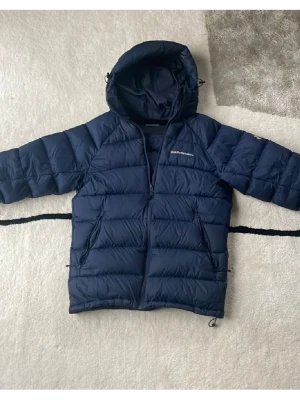 Mörkblå dunjacka från Peak Performance - Snygg mörkblå dunjacka från Peak Performance med huva och dragkedja framtill. Jackan är quiltad och har två stora fickor med dragkedja. Perfekt för kalla dagar och har en sportig look.