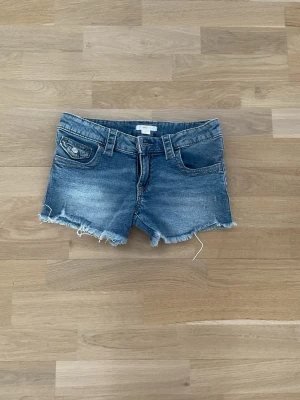 Blå jeansshorts med fransig kant - Snygga blå jeansshorts med fransig kant och klassisk femficksdesign. Shortsen har låg midja och slitna detaljer för en avslappnad look. Perfekta för varma sommardagar och enkel att matcha med olika toppar.