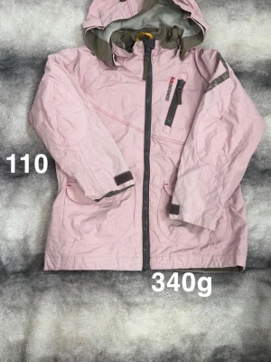 Ljusrosa vindjacka med huva - Ljusrosa vindjacka från Didriksons med huva och hel dragkedja framtill. Jackan har en bröstficka med dragkedja, två sidofickor och justerbara ärmslut med kardborre. Perfekt för blåsiga dagar och enkel att bära med sig.🌸Diffusa fläckar. Storlek 110