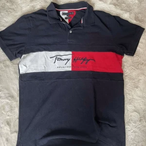 Tommy Hilfiger piké i navy, röd och vit - Snygg pikétröja från Tommy Hilfiger i slim fit med klassisk krage och korta ärmar. Tröjan är marinblå med breda ränder i rött och vitt över bröstet samt broderad logga. Tillverkad i bomull, perfekt för en clean och sportig stil.