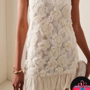 Vit blommig kortklänning med volang - Supersöt vit kortklänning med 3D-blommor över hela klänningen och volang nertill. Klänningen är ärmlös och har en romantisk känsla med sina detaljerade blommor och lätt transparent tyg. Perfekt för dig som vill sticka ut med en unik och feminin stil.