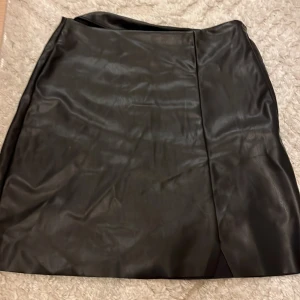 Svart skinnkjol med slits - Snygg svart kort skinnkjol med en stilren design och diskret slits framtill. Kjolen har en lätt glansig yta och stängs med dragkedja i sidan. Perfekt för dig som vill ha en edgy och trendig look.