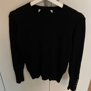 Svart långärmad tröja med knappar - En svart långärmad tröja med rund hals och snygga guldfärgade knappar vid ärmsluten. Tröjan har en klassisk passform och är perfekt för dig som gillar stilrena plagg med en liten twist. Materialet känns mjukt och passar bra till både jeans och kjol.