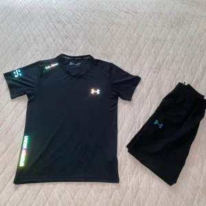 Svart Under Armour Set - Svart kortärmad t-shirt och byxor från Under Armour med reflekterande logga och textdetaljer i regnbågsfärger. Tillverkad i lätt och snabbtorkande polyester, perfekt för träning. Snygg och sportig passform.