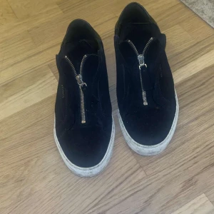 Axel arigato skor med dragkedja - Svarta sneakers i mocka från Axel Arigato med cool dragkedjedetalj framtill istället för klassisk snörning. Skorna har vit sula och en stilren, urban look som passar perfekt till jeans eller streetwear.