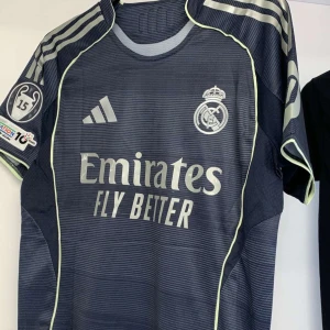 Real Madrid Huijsen matchtröja blå - Snygg Real Madrid fotbollströja från Adidas med Huijsen och nummer 24 på ryggen. Tröjan är mörkblå med silverdetaljer, tunna horisontella ränder och klubbmärke på bröstet. Korta ärmar och broderade loggor. Perfekt för dig som älskar fotboll och Real! Höst tröja