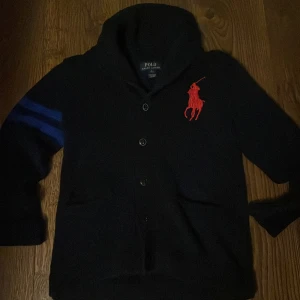 Svart kofta från Polo Ralph Lauren - Svart kofta från Polo Ralph Lauren med stora röda loggan broderad på bröstet och två blå ränder på ena ärmen. Tröjan har knäppning framtill, huva och är långärmad. Perfekt för dig som gillar klassisk men sportig stil.