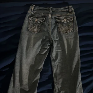 Grå bootcut jeans med broderade fickor - Snygga grå jeans med bootcut passform och broderade bakfickor med lock och knapp. Jeansen har slitna benslut och klassisk femficksdesign. Perfekta för dig som gillar detaljer och en avslappnad stil.