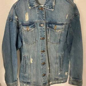 Oversized jeansjacka från Zara - Säljer en oversized ljusblå jeansjacka från Zara med slitna detaljer och klassisk krage. Jackan har knappar framtill, två bröstfickor med lock och två sidofickor. Perfekt för dig som gillar en avslappnad och trendig look.