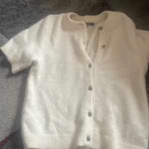 Vit kortärmad kofta från Gina Tricot - Superfin vit kofta från Gina Tricot med korta ärmar och rund hals. Koftan har ribbade muddar och knäpps framtill med silvriga knappar. Mjuk och stickad känsla, perfekt för lager-på-lager och en clean look.