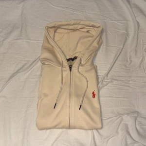 Beige hoodie från Polo Ralph Lauren - Snygg beige hoodie från Polo Ralph Lauren med dragkedja och huva. Tröjan har en röd broderad logga på bröstet, känguruficka och ribbade muddar. Tillverkad i 100% bomull för en skön och avslappnad känsla. Perfekt för chill dagar.