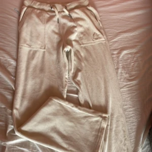Beiga mjukisbyxor Juicy Couture - Supermjuka beiga mjukisbyxor från Juicy Couture med broderad logga på fickan, snörning i midjan och stora fickor både fram och bak. Byxorna har en klassisk loose passform och är perfekta för en chill och avslappnad stil.