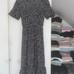 Svart prickig långklänning med volang - Svart långklänning med vita prickar och volang nertill. Klänningen har korta puffärmar och resår i midjan för en bättre passform. Knäppning med knapp i nacken.