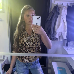 Leopardmönstrad ärmlös blus - Säljer en ärmlös blus med leopardmönster i bruna och svarta toner. Toppen har en liten krage och v-ringning, samt en lös och luftig passform. Perfekt för dig som vill sticka ut med ett djurmönster och gillar trendiga detaljer.