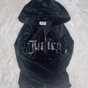 Svart velour hoodie Juicy Couture XS - Svart hoodie i mjuk velour från Juicy Couture med glittrande stenar som bildar texten 'Juicy' på bröstet. Tröjan har huva, dragkedja och långa ärmar. Perfekt för dig som gillar en klassisk Y2K-stil.