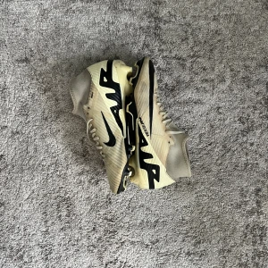 Nike Air Zoom Mercurial fotbollsskor - Snygga Nike Air Zoom Mercurial fotbollsskor i beige och svart med coola grafiska detaljer. Skorna har snörning, högre strumpliknande skaft och platt sula med dobbar för bästa grepp på planen. Lätta och smidiga i syntetmaterial, perfekta för dig som vill sticka ut på matchen.