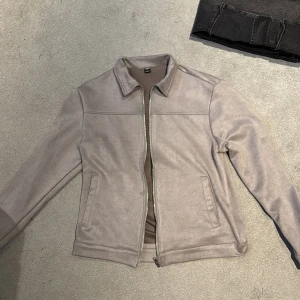 Greige mockajacka med dragkedja - Snygg greige mockajacka med klassisk krage och raka linjer. Jackan har dragkedja framtill och två diskreta fickor på sidorna. Perfekt för dig som gillar stilren och minimalistisk look.