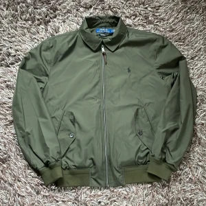 Olivgrön vindjacka från Polo Ralph Lauren - Snygg olivgrön vindjacka från Polo Ralph Lauren med klassisk krage, dragkedja med läderdetalj och broderad logga på bröstet. Jackan har ribbade muddar, två fickor med knapp och är perfekt för dig som gillar stilren och sportig look.