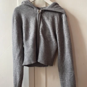 Grå stickad hoodie med dragkedja - Grå stickad hoodie från Gina Tricot med dragkedja framtill. Tröjan är croppad med ribbade muddar och midja, har huva och långa ärmar. Perfekt för lager-på-lager och chill dagar.