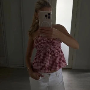 Rutig röd tubtopp från Stradivarius - Supersöt röd och vit rutig tubtopp från Stradivarius. Toppen är bandeau-modell med smockad midja och volangig nederdel. Perfekt till vita jeans eller shorts för en somrig look. Gjord i mjuk bomull som känns skön mot huden.