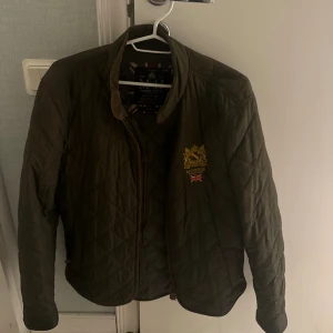 Olivgrön quiltad jacka från Morris - Snygg olivgrön quiltad jacka från Morris med guldbroderad logga och brittisk flagga på bröstet. Jackan har ståkrage, dragkedja och klassiskt rutmönster. Perfekt för dig som gillar preppy stil och vill ha något som sticker ut.