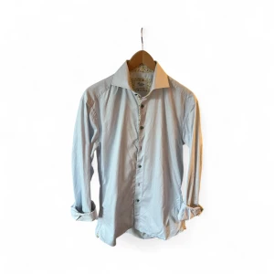 Ljusblå randig skjorta från Stenströms - Snygg ljusblå skjorta från Stenströms med diskreta ränder och coola gröna knappar. Insidan av kragen och knappslån har ett blommigt mönster i gult och rött. Skjortan är i bomull och har lång ärm. Perfekt för dig som gillar detaljer och stilren look.