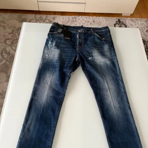 Dsquared2 blå jeans med slitningar - Snygga blå jeans från Dsquared2 med coola slitningar och färgstänk. Klassisk femficksmodell med orange sömmar och läderpatch bak. Jeansen har en rak passform och är tillverkade i bomull. Perfekta för dig som gillar en streetig och trendig look.