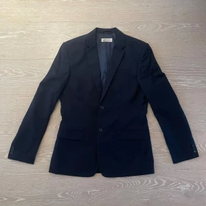 Mörkblå kavaj från H&M, storlek 170 - Snygg mörkblå kavaj från H&M i klassisk modell med två knappar framtill och fickor med lock. Kavajen har smal krage och är helfodrad med slät insida. 