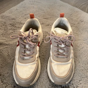 Beige sneakers från K.Cobler strl 38 - Snygga chunky sneakers från K.Cobler i beige och grått med röda detaljer på sidorna. Skorna har snörning med vita och röda skosnören, samt orange hälflik. Ovandel i textil och syntet, sula i phylon och TPR. Perfekta för en trendig streetstyle-look.