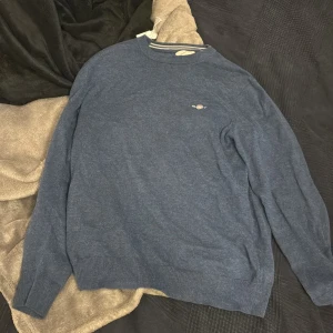 Blå tröja med broderad detalj - Mysig blå tröja med rund hals och diskret broderad logga på bröstet. Tröjan har långa ärmar och ribbade muddar vid ärmslut och nederkant. Perfekt för lager-på-lager och enkel att matcha med jeans eller chinos.