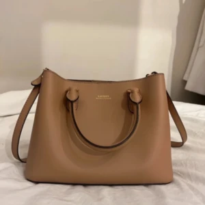 Beige handväska från Lauren Ralph Lauren - Snygg beige handväska från Lauren Ralph Lauren i stilren design. Väskan har både handtag och axelrem, guldiga metalldetaljer och flera fack inuti, inklusive ett med dragkedja. Tillverkad i slätt skinn som ger en lyxig känsla.