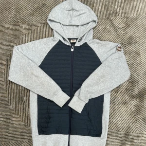 Colmar cardigan  - Snygg hoodie från Colmar Originals i grått och svart. Tröjan har quiltad svart front och rygg, grå ärmar och huva samt dragkedja hela vägen. Perfekt för dig som gillar sportig och modern stil. Colmar-logga på ärmen ger en extra touch.