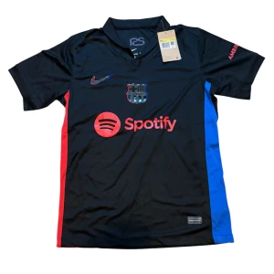 Svart FC Barcelona fotbollströja Nike - Snygg svart FC Barcelona fotbollströja från Nike med röda och blå detaljer på sidorna. Tröjan har klubbmärke på bröstet, Spotify-logga framtill och UNHCR-tryck på ryggen. Tillverkad i lätt och ventilerande polyester, perfekt för match eller träning.