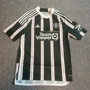 Manchester United kit away 2022/23 - Snygg Manchester United fotbollströja från Adidas med gröna och vita vertikala ränder och röda detaljer. Tröjan har korta ärmar, vit krage och klassiska Adidas-ränder på axlarna. Tillverkad i lätt och ventilerande polyester med TeamViewer-logga på bröstet.
