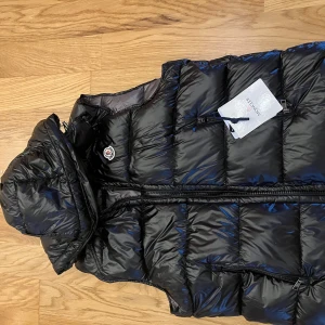 Svart dunväst från Moncler med luva - Säljer en svart, glansig dunväst från Moncler med avtagbar luva och dragkedja framtill. Västen har quiltad design, två fickor med dragkedja och Moncler-logga på bröstet. Perfekt för lager-på-lager och snygg till streetwear.