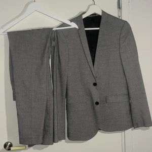 Grå kostym med kavaj och byxor - Snygg grå kostym med klassisk kavaj och matchande byxor. Kavajen har två knappar framtill, smal krage och fickor med lock. Byxorna är raka och har bälteshällor. Materialet är mjukt och har en lätt struktur som ger en stilren look.