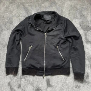 Svart jacka Limitato x Riocam - Svart jacka med coolt konstmotiv på ryggen i starka färger. Jackan har silverfärgade dragkedjor, två snedställda fickor och broderad Limitato-logga framtill. Mjuk bomullskänsla och klassisk krage ger en clean look.