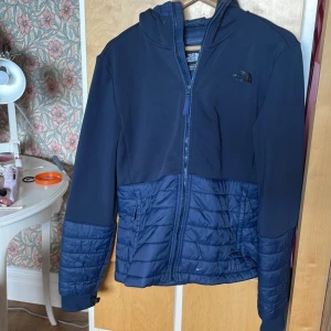 Blå hybridjacka från The North Face - Snygg blå hybridjacka från The North Face med huva och hel dragkedja. Jackan har quiltade partier i glansigt material på nedre delen och ärmarna, samt slät ovandel. Perfekt för dig som vill ha en sportig och stilren look. Fickor med dragkedja på sidorna.