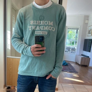 Tröja Morris - Snygg grön långärmad tröja från Morris Company med tryck framtill och ankare som detalj. Tröjan har ribbade muddar vid ärmslut och nederkant, och är tillverkad i ett mjukt bomullsmaterial. Perfekt för dig som gillar stilren och avslappnad look. Perfekt för höst och vinter